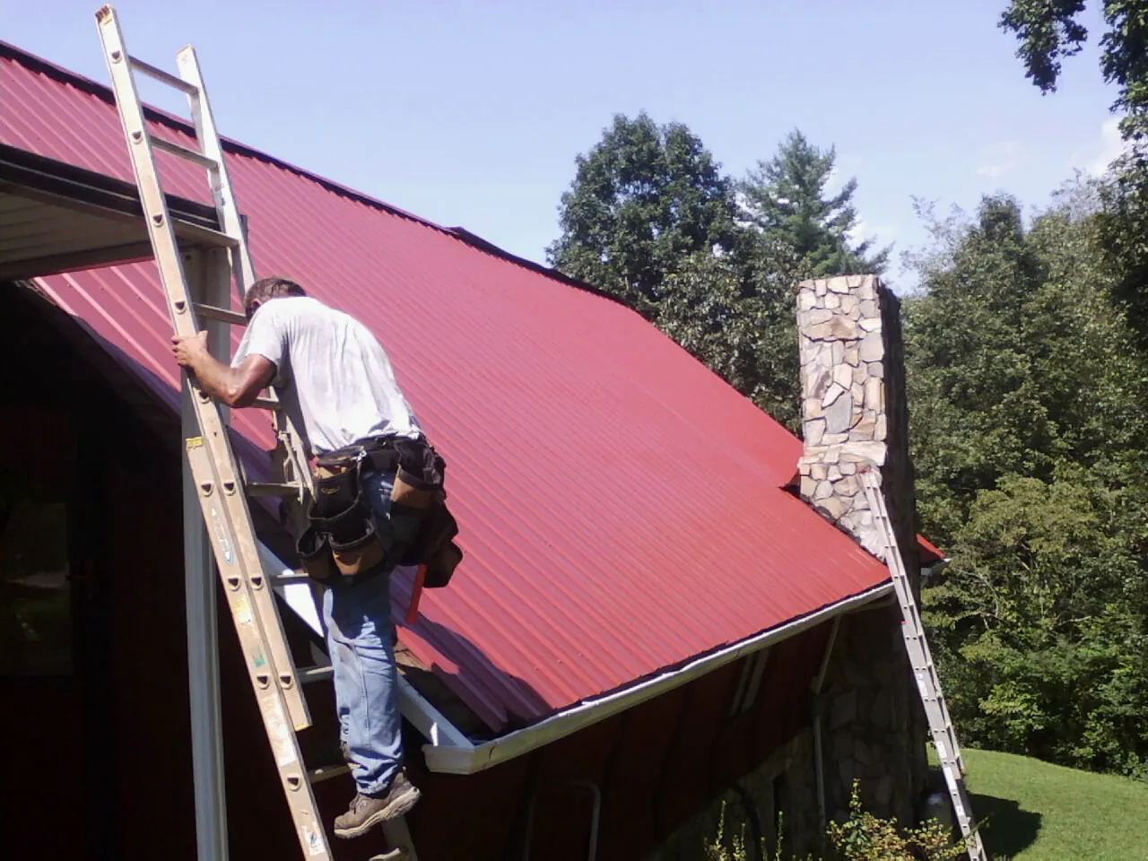 Roofer on a ladder beside a red metal roof and stone chimney — Soffit & Fascia Repair by Roofing Brothers in West Hartford